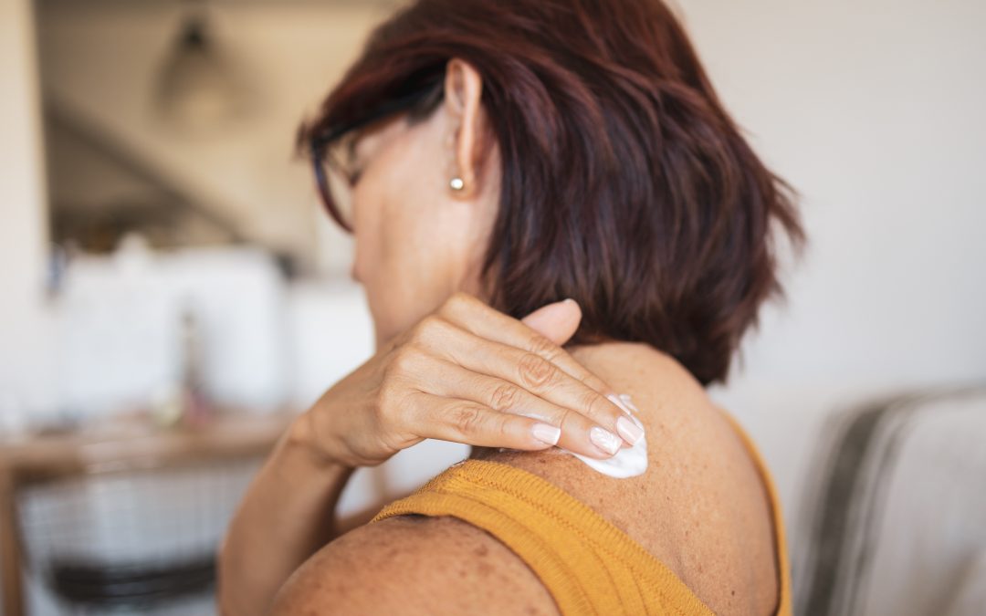 A senior Latin woman is sitting on her living room sofa and applying a hormone cream to her back